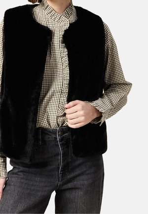 Persoon draagt een zwart nepbontvest over een beige en zwart geruite blouse met ruches en donkergrijze jeans met hoge taille, hand rust dicht bij de taille.