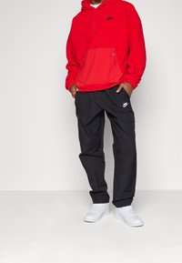 Felpa rossa con zip frontale e tasca, logo nero, abbinata a pantaloni cargo neri e sneaker bianche. Tessuto spesso, vestibilità comoda.