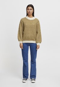 Pull en tricot à motifs en losange marron et crème, avec un col rond et des manches bouffantes ; associé à un jean bleu évasé et des baskets noires.