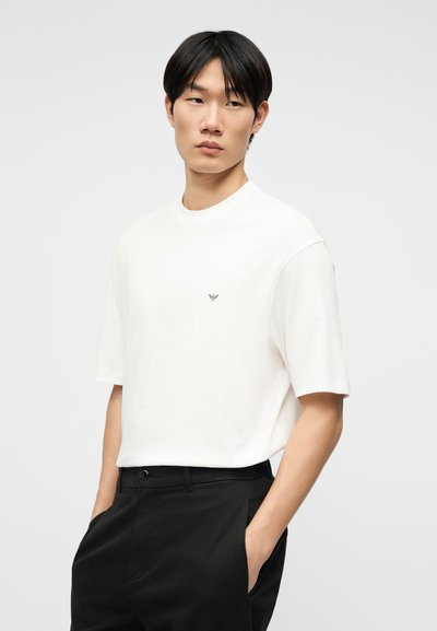 Νεαρός άνδρας που φοράει λευκό πουκάμισο με κοντά μανίκια και μικρό λογότυπο στο στήθος και μαύρο παντελόνι, με τα χέρια στις τσέπες.