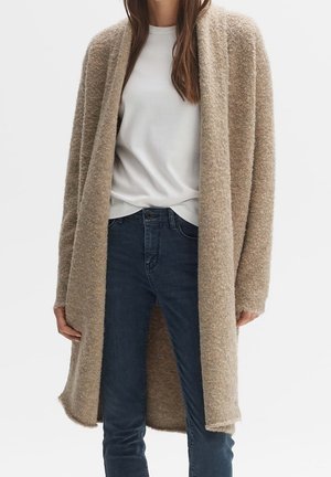 Femme portant un long cardigan beige texturé par-dessus un haut blanc et un jean bleu foncé sur un fond clair uni.