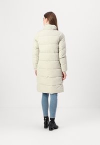 Padded long coat in light gray with a high collar, horizontal stitching, and long sleeves, paired with fitted jeans and ankle boots.