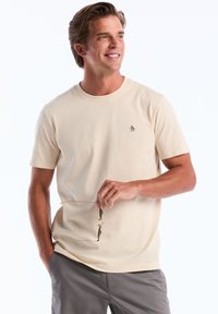 T-shirt crème à manches courtes en tissu doux, avec un petit logo de pingouin bleu marine sur la poitrine, associé à un short gris.