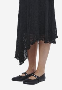 Zwarte kanten jurk met een asymmetrische zoom, gecombineerd met zwarte leren platte schoenen met decoratieve riempjes en studs.