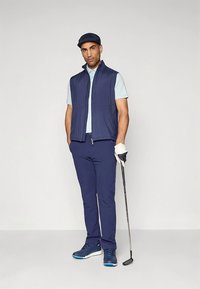 Temno modra golf obleka, ki vključuje brezrokavnik, svetlo modro polo majico, ožene hlače, temno modre čevlje in kapo, z golf palico v roki.