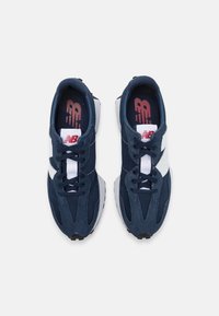 Ett par marinblå och vita New Balance-sneakers med snören, sedd uppifrån mot en ljus bakgrund.