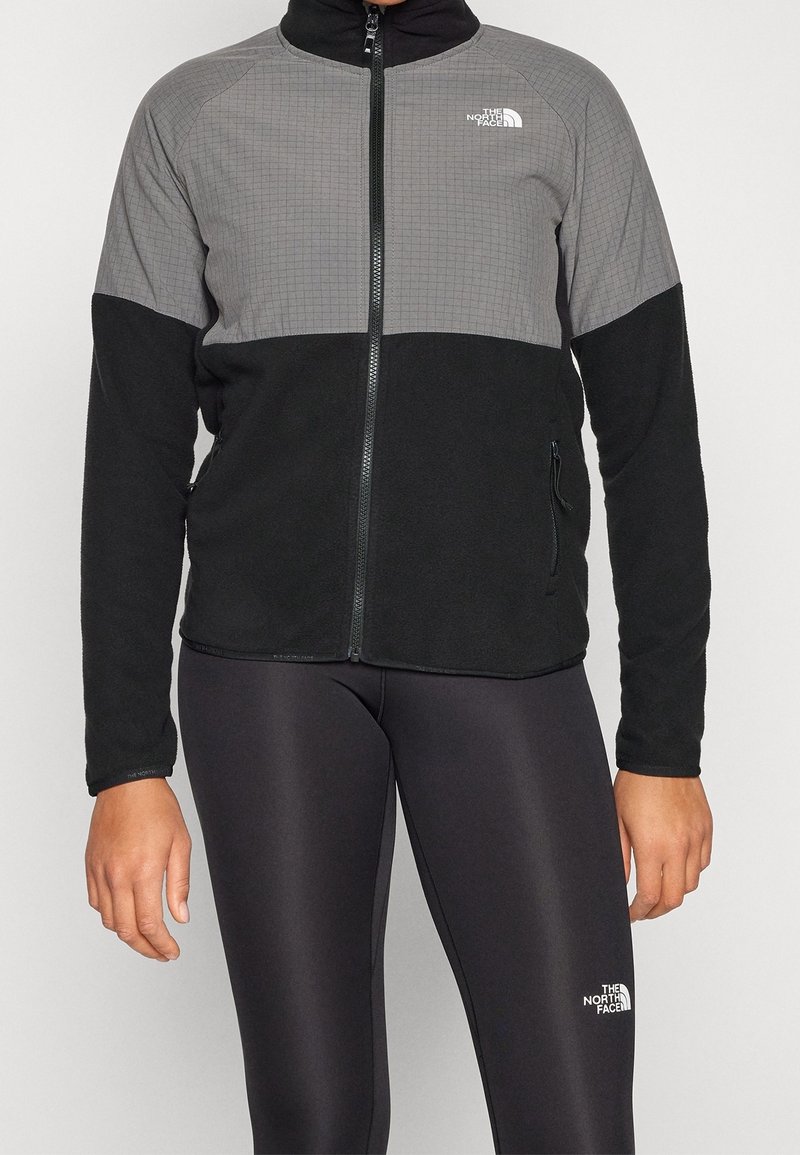 Personne portant une veste polaire à fermeture éclair noire et grise et des leggings noirs, tous deux avec des logos The North Face.