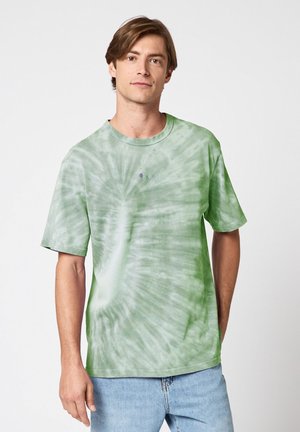 Jeune homme portant un T-shirt tie-dye vert et blanc et un jean bleu clair, debout devant un fond blanc uni.