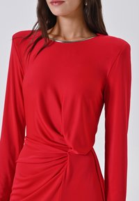 Rode jurk met lange mouwen, een gedraaide zijkant en een metalen randje langs de halslijn, gemaakt van soepel, rekbaar materiaal.