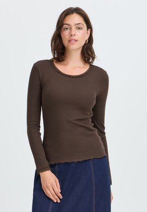 Kvinde med kort brunt hår iført en tætsiddende brun bluse med langærmede og bølget kant samt et blåt skørt, stående mod en hvid baggrund.