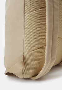 Un sac à dos beige avec un panneau en mesh et des côtés rembourrés ; fabriqué en tissu texturé avec des coutures plates et une boucle latérale pour le transport.