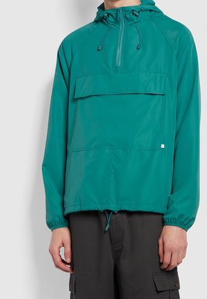 Türkis hættejakke med lynlås foran, stor klaplomme, elastiske manchetter og snoretræk i kanten, båret med sorte cargobukser.