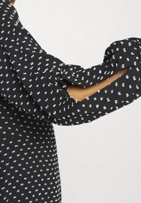 Černá tkanina s bílým puntíkovaným vzorem; má splývavý design rukávů s volány a lehkou texturu, která zvýrazňuje splývavost oděvu.