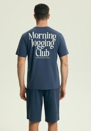 Mężczyzna w granatowych szortach i pasującej koszulce z napisem „Morning Jogging Club” stoi tyłem na jednolitym tle.