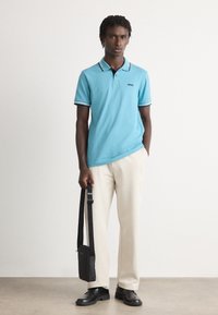 Άνδρας φορώντας ανοιχτό μπλε πόλο και μπεζ παντελόνι, κρατώντας μια μαύρη τσάντα ώμου, στέκεται μπροστά από ένα απλό φωτεινό φόντο.