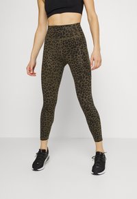 Legginsy z wysokim stanem w kolorze oliwkowym w czarno-leopardzi wzór. Charakteryzują się elastycznym materiałem i gładkim wykończeniem, w zestawieniu z czarnymi butami sportowymi.