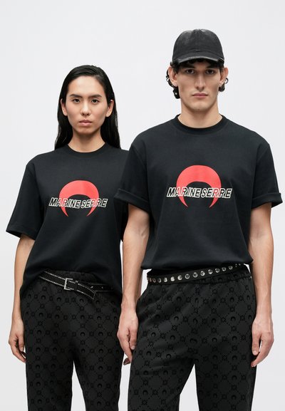 T-shirts de algodão pretas com logótipo circular vermelho, combinadas com calças pretas estampadas que apresentam um cinto de tachas metálicas e um design subtil em relevo.
