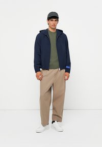 Marineblauwe lichte jas met capuchon, groene trui eronder, beige wijde broek en witte sneakers, gedragen door een mannelijke figuur. Inclusief een grijze pet.