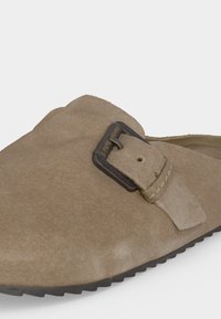 Primer plano de un zapato tipo mocasín de ante beige con hebilla de metal oscura y costuras visibles. La suela es negra con borde texturizado.
