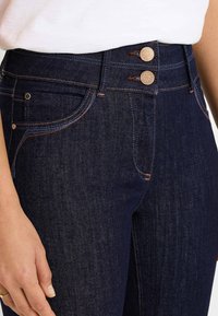 Jeans de mezclilla azul oscuro con cintura alta, cierre de dos botones, costuras en contraste y bolsillos frontales con textura suave.