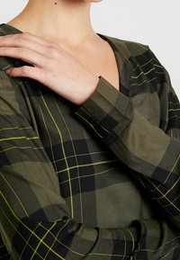 Langärmliges Hemd aus olivgrünem Stoff mit einem schwarz-gelben Karomuster, ausgestattet mit einem V-Ausschnitt und knöpfbaren Manschettendetails.