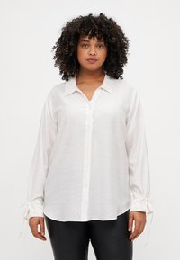 Camicia bianca a maniche lunghe con chiusura a bottoni, morbida al tatto e con dettagli a cravatta sui polsini. Presenta un colletto classico e una vestibilità rilassata.