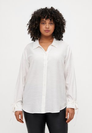 Chemise blanche à manches longues avec un devant boutonné, texture douce et accents de nœud aux poignets. Comprend un col classique et une coupe décontractée.