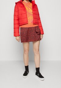 Kvinna som bär en kortad röd pufferjacka, en orange mönstrad tröja, en minikjol med röd och svart hjärtprint, svarta strumpor och svarta slip-on skor.