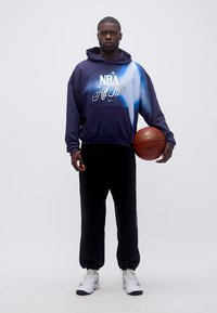 Άνδρας που φοράει μπλε σκούρο και λευκό φούτερ NBA All Star, μαύρο παντελόνι, λευκά αθλητικά παπούτσια, κρατώντας μια μπάλα μπάσκετ Spalding στο δεξί χέρι.