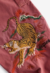 Broderad tigerdesign på rosa tyg med gröna och orange blommönster; innehåller texten "Super" i svart. Detaljerad textur och livliga färger.