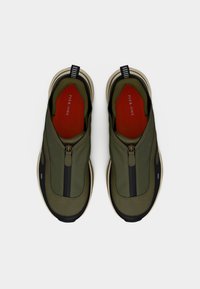 Πράσινα παπούτσια slip-on με φερμουάρ μπροστά, μαύρες λεπτομέρειες, υφή στο πάνω μέρος και κόκκινη εσωτερική σόλα σε αντίθεση. Διαθέτουν επενδεδυμένο κολάρο και λαστιχένια σόλα.