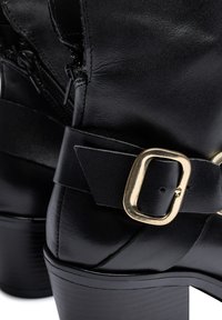 Zwarte leren enkellaars met een glanzende afwerking, voorzien van een brede zwarte band met een gouden gespsluiting en een gestructureerde blokhak.