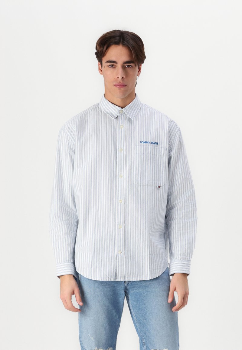 Camisa de manguitos largos a rayas en azul y blanco con cuello, que presenta un bolsillo en el pecho y el logo "TOMMY JEANS" en bordado azul.