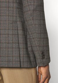 Braun-kariertes Sakko mit einem strukturierten Muster, ausgestattet mit einem Schlitz auf der Rückseite und einer Drei-Knopf-Leiste am Ärmel. Der Stoff ist gewebt und hat ein strukturiertes Design.