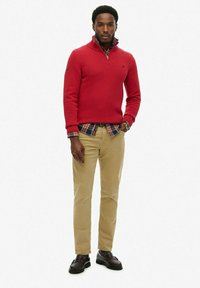 Suéter de punto rojo con cuello de cremallera sobre una camisa de cuadros, combinado con pantalones caquis y zapatos marrón oscuro. Hombre de pie con los brazos relajados.