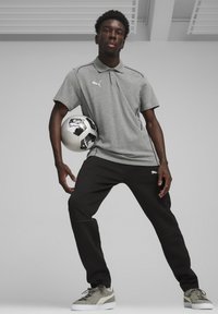 Grijze polo met korte mouwen, zwarte joggingbroek en grijze sneakers. Speler houdt een zwart-witte voetbal vast, met logo-details.