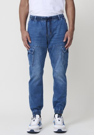 Pantalones cargo de mezclilla azul con cintura elástica, cordón y tobillos ajustados. Incluye dos grandes bolsillos laterales y un ajuste relajado.