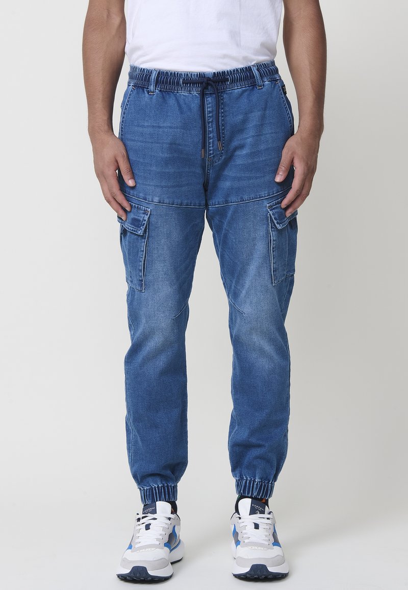 Modre jeans hlače s cargo slogom in elastičnim pasom, z vrvico in prilegajočimi se gležnji. Imajo dve veliki stranski žep in sproščen kroj.