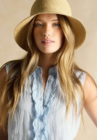 Bluză fără mâneci de un albastru deschis, cu detalii volănașe pe partea din față, asociată cu o pălărie din paie cu boruri largi. Fundal neutru.