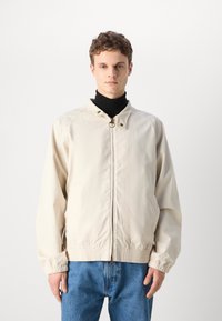 Helle beige Jacke mit Frontreißverschluss, hohem Kragen und elastischen Bündchen. Glatte Textur, mit Raglanärmeln und minimalistischem Design.