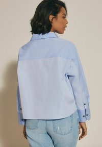 Cămașă oversized de culoare albastru deschis cu un design cu despicătură pe spate, având mâneci lungi și accente cu nasturi negri. Purtați-o cu blugi albaștri decolorati.