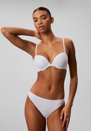Femme posant en soutien-gorge blanc et sous-vêtements assortis sur un fond gris uni, un bras levé derrière la tête.
