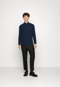 Chemise à manches longues boutonnée de couleur marine avec poche, associée à un pantalon noir et des chaussures, sur fond blanc uni.