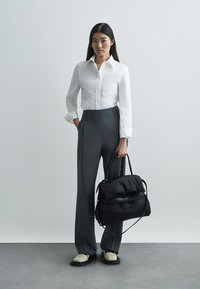 Fehér vászon gombos ing, magas derekú szürke nadrág, fekete párnázott táska és krémszínű cipők egy minimalista környezetben.