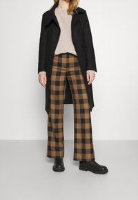 Sort dobbeltradet frakke, beige sweater og brune bukser i ternet mønster med brede ben. Sorte klodsede støvler fuldender outfittet. Neutral baggrund.