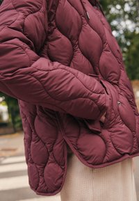 Doudoune matelassée bordeaux avec une texture ondulée, capuche et poches latérales avec des accents de ferrures noires. Superposition en beige côtelé en dessous.