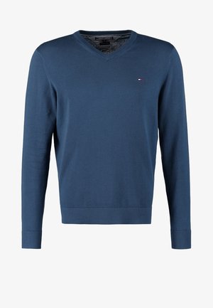 Pull bleu à col en V, maille légère ; jean foncé coupe slim ; chaussures noires ; portant un sac messager en cuir marron.