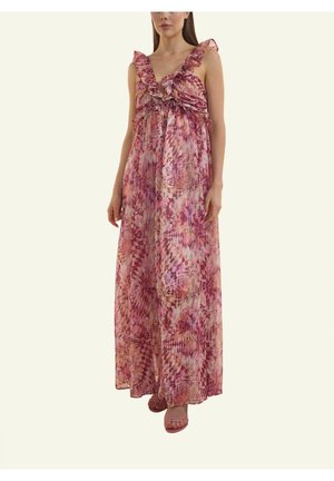 Woman wearing sleeveless pink patterned maxi dress with ruffled V-neck and strappy pink sandals, standing against plain background.