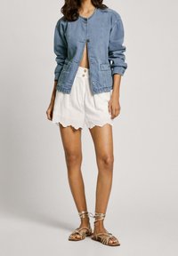 Chaqueta de mezclilla azul claro con cuello redondo, presenta cierre de botones, combinada con unos pantalones cortos blancos con dobladillo en forma de ondas y sandalias doradas.