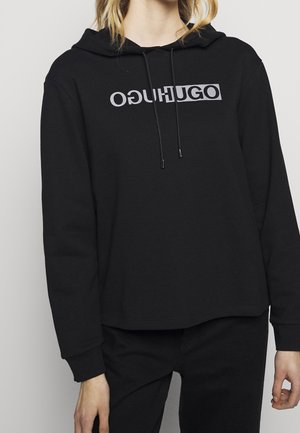 Person trägt einen schwarzen Hoodie mit dem "HUGO"-Logo, das auf der Brust sowohl spiegelverkehrt als auch normal gedruckt ist, kombiniert mit einer schwarzen Hose.